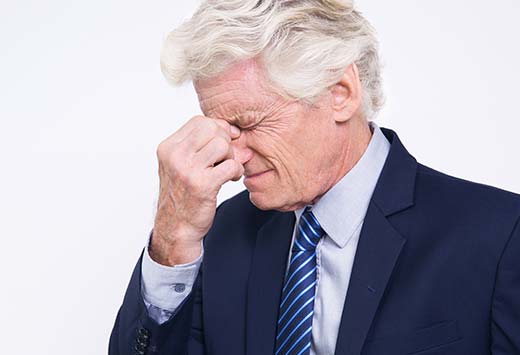 older man in a suit pinching the bridge of his nose as if he's in pain